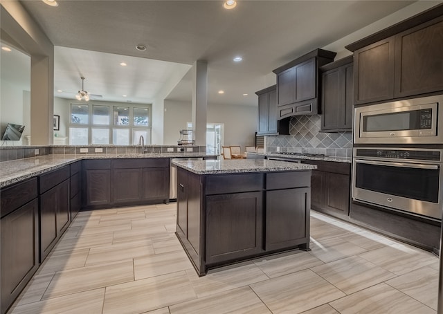 kitchen featuring decorative backsplash, light stone counters, dark brown cabinets, stainless steel appliances, and ceiling fan