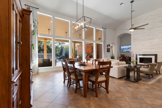 dining area featuring ceiling fan with notable chandelier, light tile patterned floors, a fireplace, and high vaulted ceiling