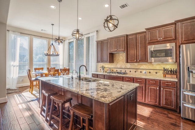 kitchen with sink, hanging light fixtures, dark wood-type flooring, stainless steel appliances, and a center island with sink