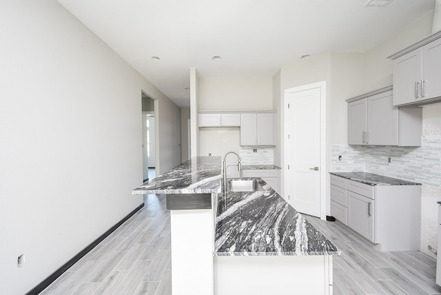 kitchen featuring dark stone counters, a center island with sink, sink, light hardwood / wood-style flooring, and tasteful backsplash