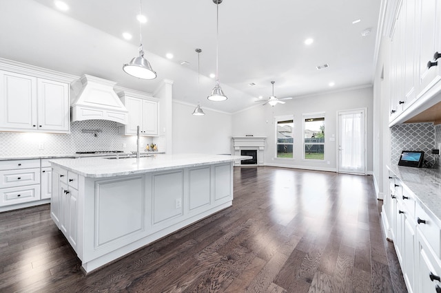 kitchen with backsplash, premium range hood, dark wood-type flooring, an island with sink, and decorative light fixtures