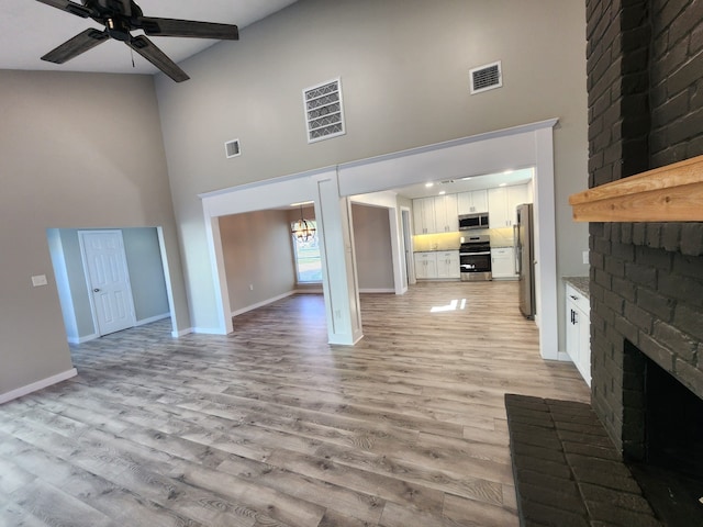 unfurnished living room with ceiling fan with notable chandelier, light hardwood / wood-style flooring, high vaulted ceiling, and a brick fireplace