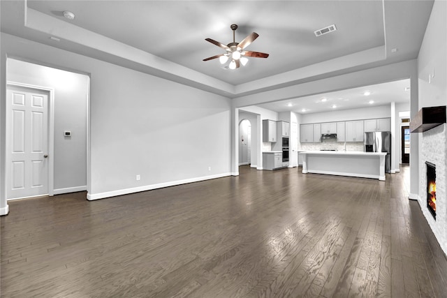 unfurnished living room featuring a raised ceiling, a stone fireplace, ceiling fan, and dark hardwood / wood-style flooring