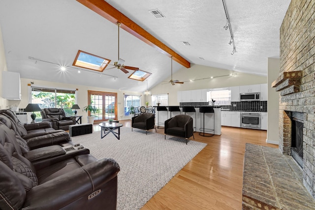 living room with ceiling fan, a brick fireplace, light hardwood / wood-style flooring, a textured ceiling, and vaulted ceiling with skylight