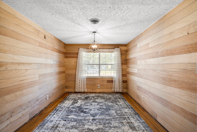 unfurnished dining area with a chandelier, a textured ceiling, dark hardwood / wood-style flooring, and wooden walls