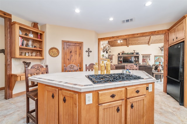 kitchen with beamed ceiling, tile countertops, stainless steel gas stovetop, black refrigerator, and a kitchen island