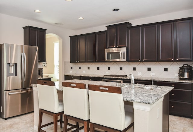 kitchen with light stone countertops, sink, stainless steel appliances, tasteful backsplash, and an island with sink