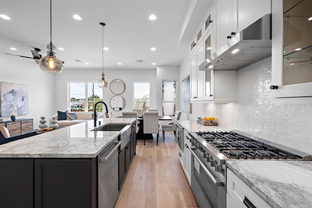 kitchen featuring appliances with stainless steel finishes, a kitchen island with sink, decorative light fixtures, white cabinetry, and range hood