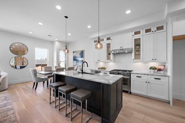 kitchen featuring light wood-type flooring, high end stove, pendant lighting, white cabinets, and an island with sink