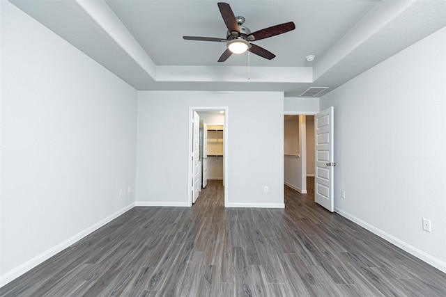 unfurnished bedroom with ceiling fan, dark wood-type flooring, a tray ceiling, a walk in closet, and a closet