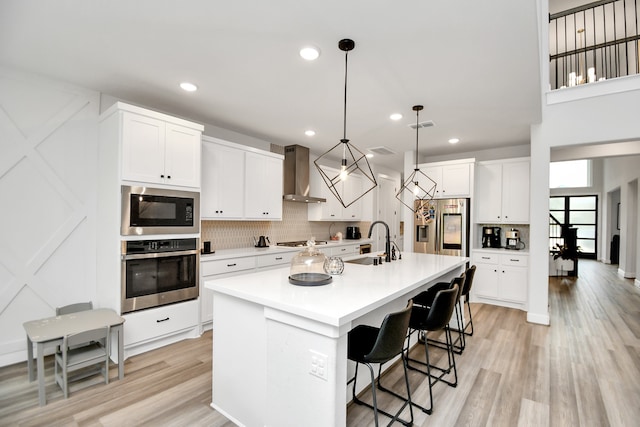 kitchen with a kitchen breakfast bar, wall chimney range hood, hanging light fixtures, an island with sink, and appliances with stainless steel finishes