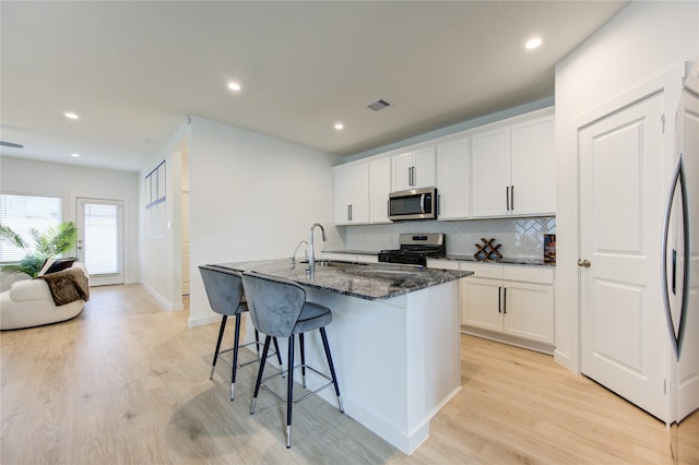 kitchen featuring light hardwood / wood-style flooring, dark stone countertops, an island with sink, appliances with stainless steel finishes, and white cabinetry