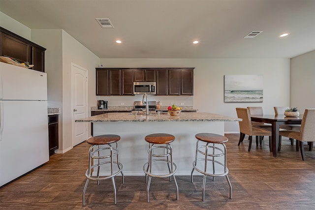 kitchen featuring dark wood-type flooring, an island with sink, appliances with stainless steel finishes, light stone counters, and dark brown cabinetry