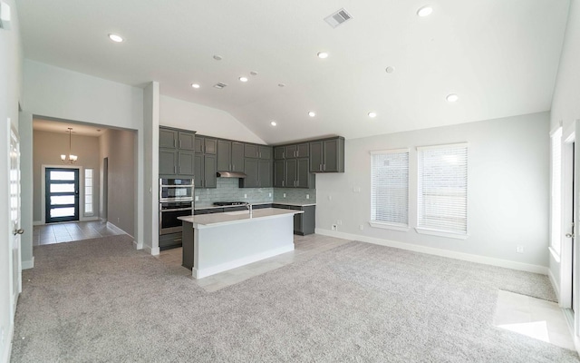 kitchen featuring vaulted ceiling, double oven, light colored carpet, a kitchen island with sink, and backsplash