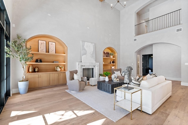 living room with an inviting chandelier, a towering ceiling, light hardwood / wood-style flooring, a premium fireplace, and built in shelves