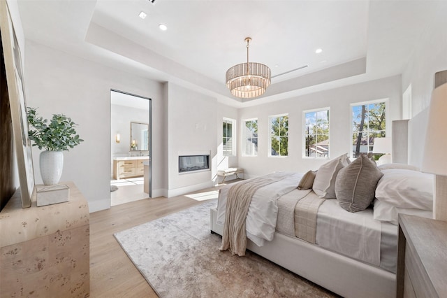 bedroom with a raised ceiling, an inviting chandelier, connected bathroom, and light hardwood / wood-style flooring