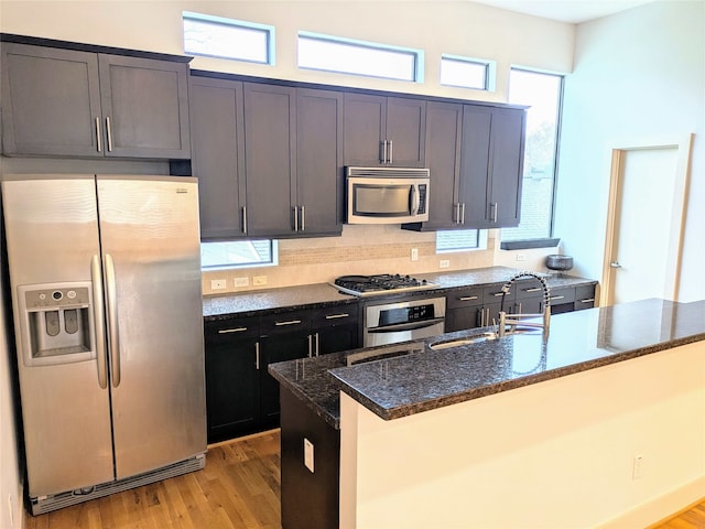 kitchen with dark stone countertops, a kitchen island with sink, stainless steel appliances, light wood-type flooring, and sink