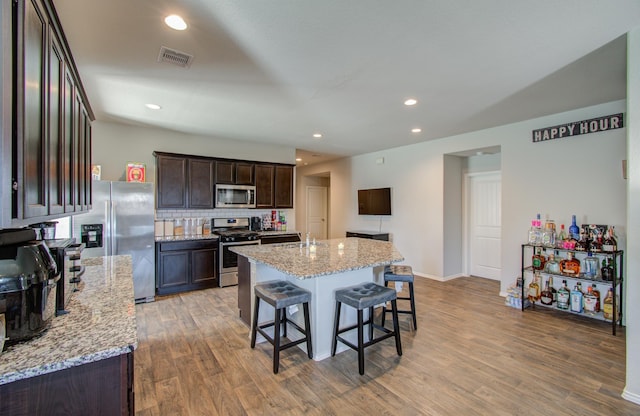 kitchen with hardwood / wood-style floors, stainless steel appliances, light stone counters, and a center island with sink