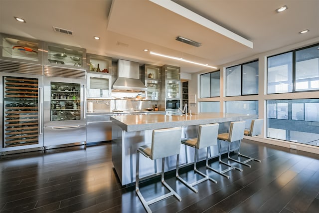 kitchen with dark wood-type flooring, a center island with sink, a kitchen breakfast bar, wine cooler, and wall chimney exhaust hood