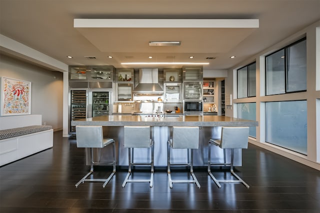 kitchen featuring a spacious island, wall chimney exhaust hood, dark hardwood / wood-style flooring, and a breakfast bar