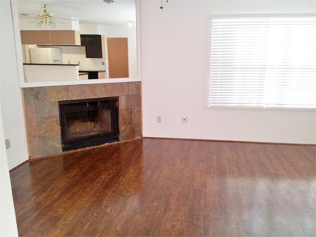 unfurnished living room featuring ceiling fan, a healthy amount of sunlight, dark wood-type flooring, and a tiled fireplace