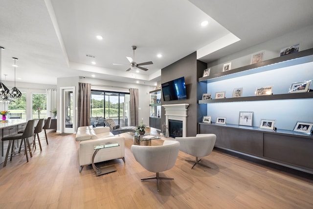 living room featuring ceiling fan with notable chandelier, a raised ceiling, and light hardwood / wood-style flooring