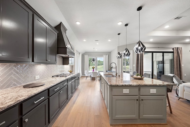 kitchen featuring premium range hood, a kitchen island with sink, sink, pendant lighting, and light hardwood / wood-style flooring