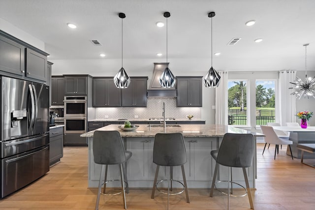 kitchen featuring a center island with sink, hanging light fixtures, light hardwood / wood-style flooring, light stone countertops, and appliances with stainless steel finishes