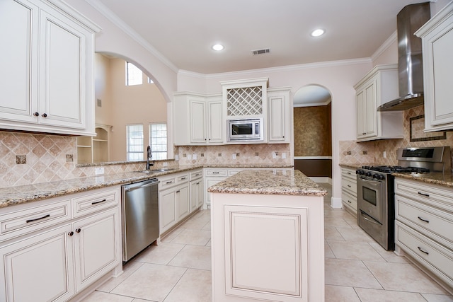 kitchen featuring wall chimney range hood, ornamental molding, a kitchen island, light stone counters, and stainless steel appliances