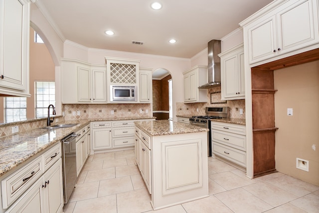 kitchen with wall chimney range hood, sink, light stone countertops, appliances with stainless steel finishes, and a kitchen island