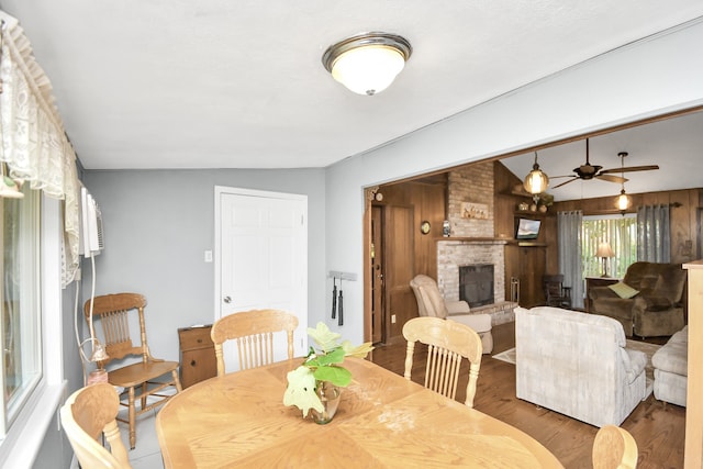 dining area with ceiling fan, a brick fireplace, wood-type flooring, vaulted ceiling, and wooden walls