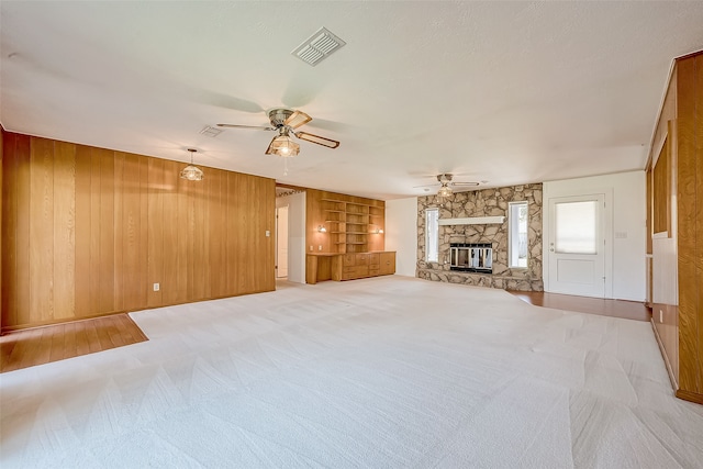 unfurnished living room with ceiling fan, a stone fireplace, built in features, wood walls, and light carpet