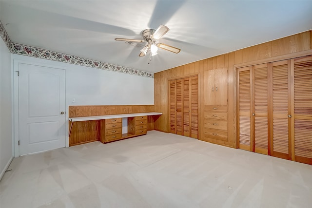 unfurnished bedroom with light colored carpet, ceiling fan, built in desk, multiple closets, and wood walls