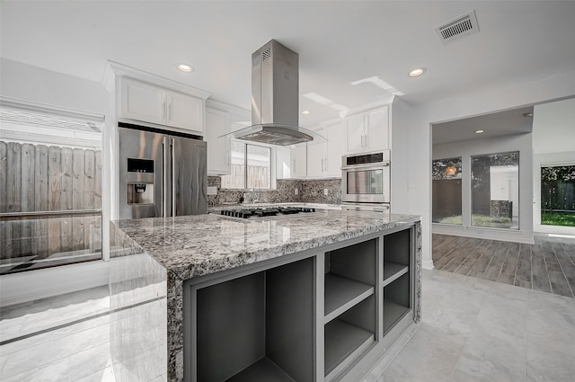 kitchen with island exhaust hood, appliances with stainless steel finishes, light stone countertops, tasteful backsplash, and white cabinets