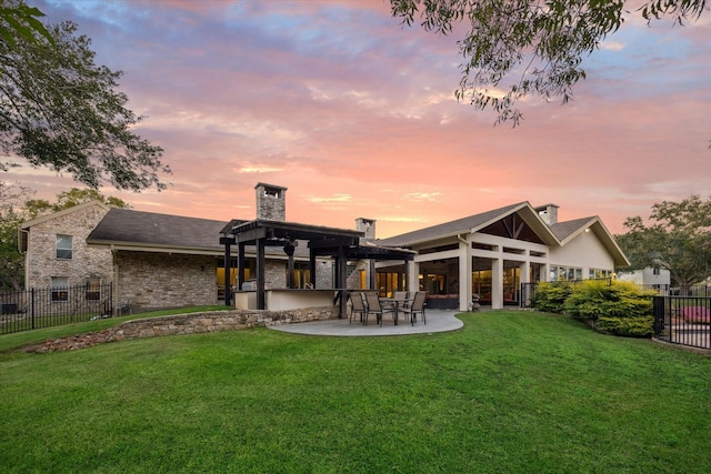 back of house at dusk featuring fence, stone siding, a lawn, a chimney, and a patio area