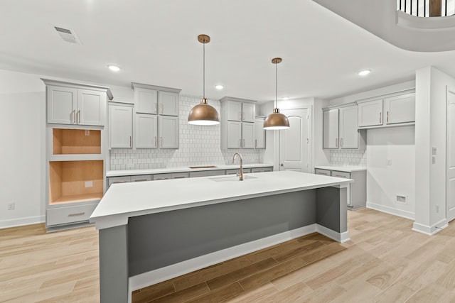 kitchen featuring sink, tasteful backsplash, gray cabinets, a kitchen island with sink, and light wood-type flooring