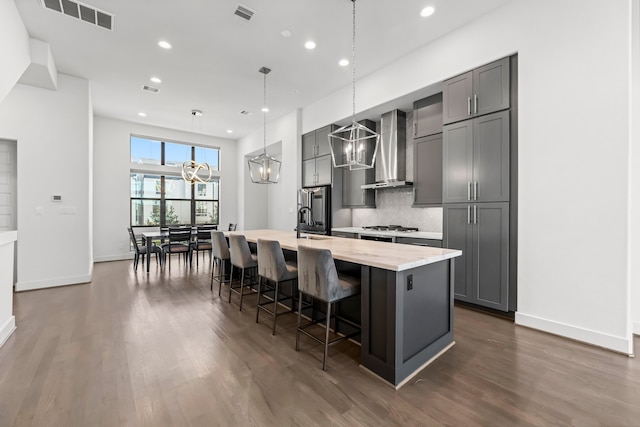 kitchen with wall chimney range hood, dark hardwood / wood-style floors, decorative light fixtures, gray cabinets, and a center island with sink