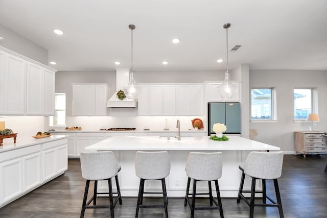 kitchen with stainless steel refrigerator, a center island with sink, pendant lighting, and custom range hood