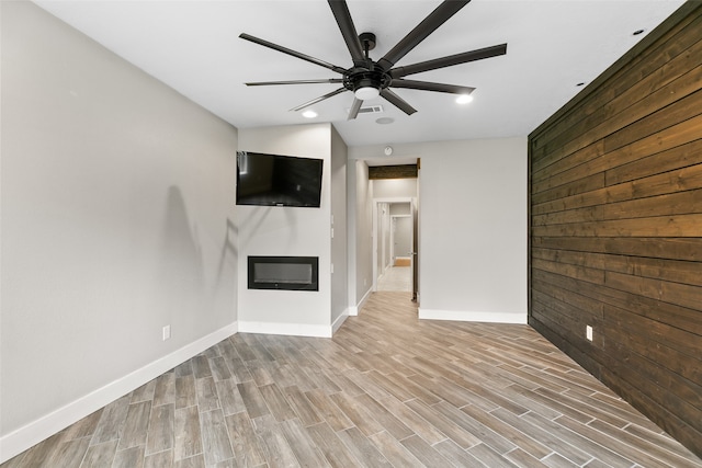 unfurnished living room featuring ceiling fan, light wood-style flooring, wood walls, baseboards, and a glass covered fireplace