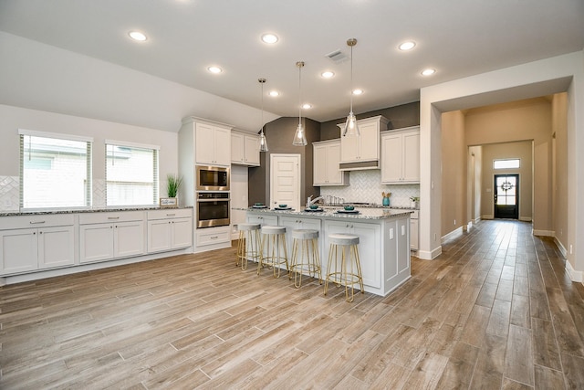 kitchen with appliances with stainless steel finishes, white cabinets, light hardwood / wood-style floors, hanging light fixtures, and an island with sink