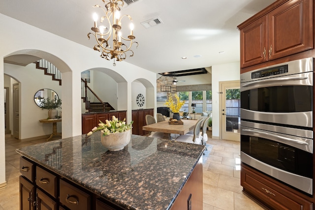 kitchen with dark stone counters, ceiling fan with notable chandelier, stainless steel double oven, and decorative light fixtures