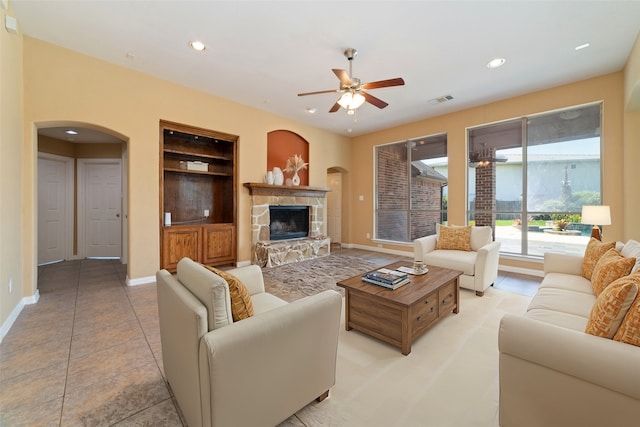 living room with a stone fireplace, ceiling fan, built in shelves, and light tile patterned floors