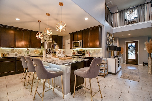 kitchen featuring light stone countertops, appliances with stainless steel finishes, backsplash, pendant lighting, and a center island with sink