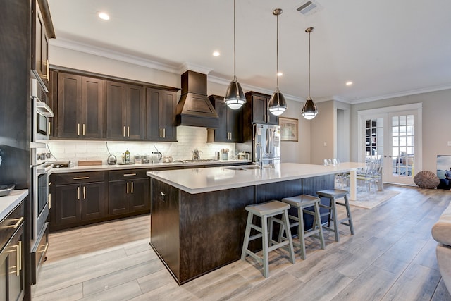 kitchen with custom exhaust hood, decorative light fixtures, light hardwood / wood-style flooring, an island with sink, and dark brown cabinets