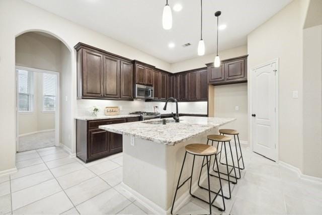 kitchen featuring a kitchen island with sink, sink, hanging light fixtures, light stone countertops, and dark brown cabinets