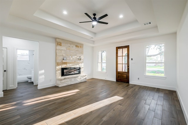 unfurnished living room with a tray ceiling, a stone fireplace, ceiling fan, and dark wood-type flooring