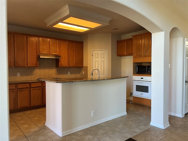 kitchen with white oven, stone countertops, an island with sink, and backsplash