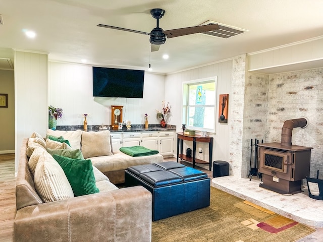 living room featuring wood-type flooring, a wood stove, ceiling fan, and crown molding