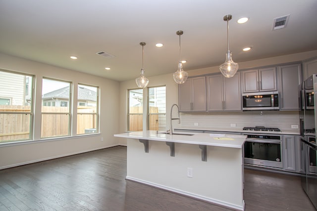 kitchen with appliances with stainless steel finishes, sink, pendant lighting, dark hardwood / wood-style floors, and an island with sink