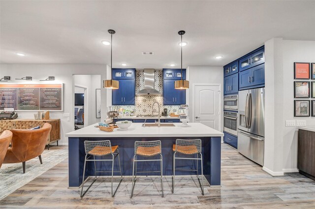 kitchen with wall chimney range hood, light hardwood / wood-style flooring, blue cabinets, decorative light fixtures, and appliances with stainless steel finishes